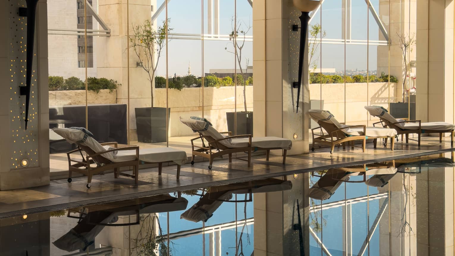 A smooth, serene indoor pool surrounded by lounge chairs, with floor-to-ceiling windows admitting natural light and outdoor views