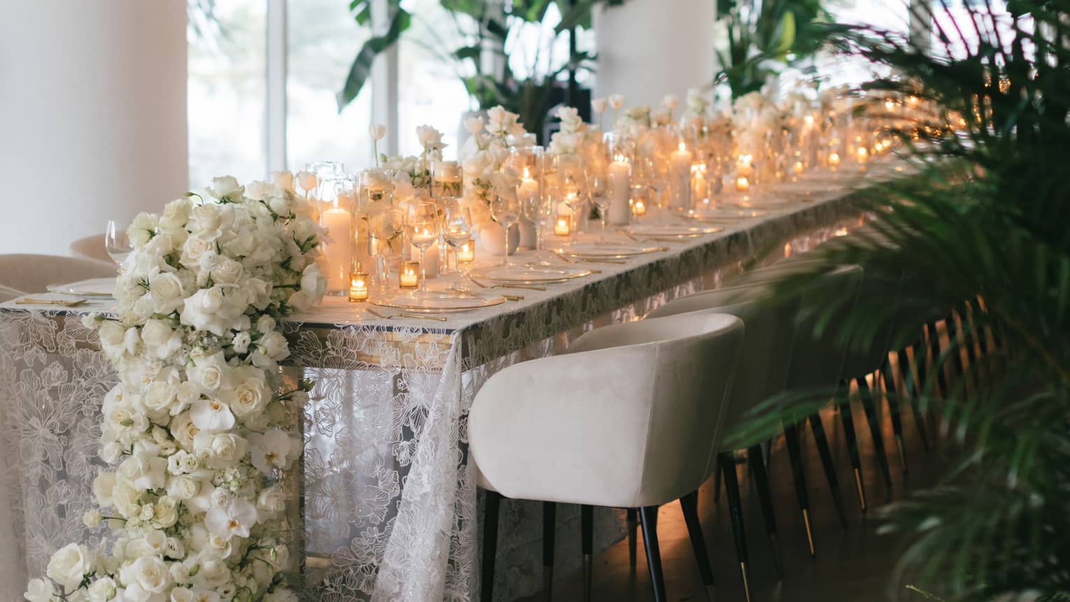 Long rectangular table topped with a white lace tablecloth and a variety of votive candles and white floral garlands lining the centre