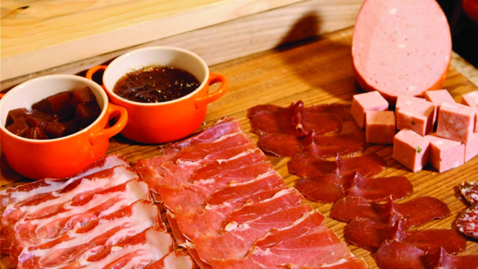 Rows of finely sliced red meats and chunks of cold cuts laid out on a wood board, before a leg of meat held by a metal clamp.