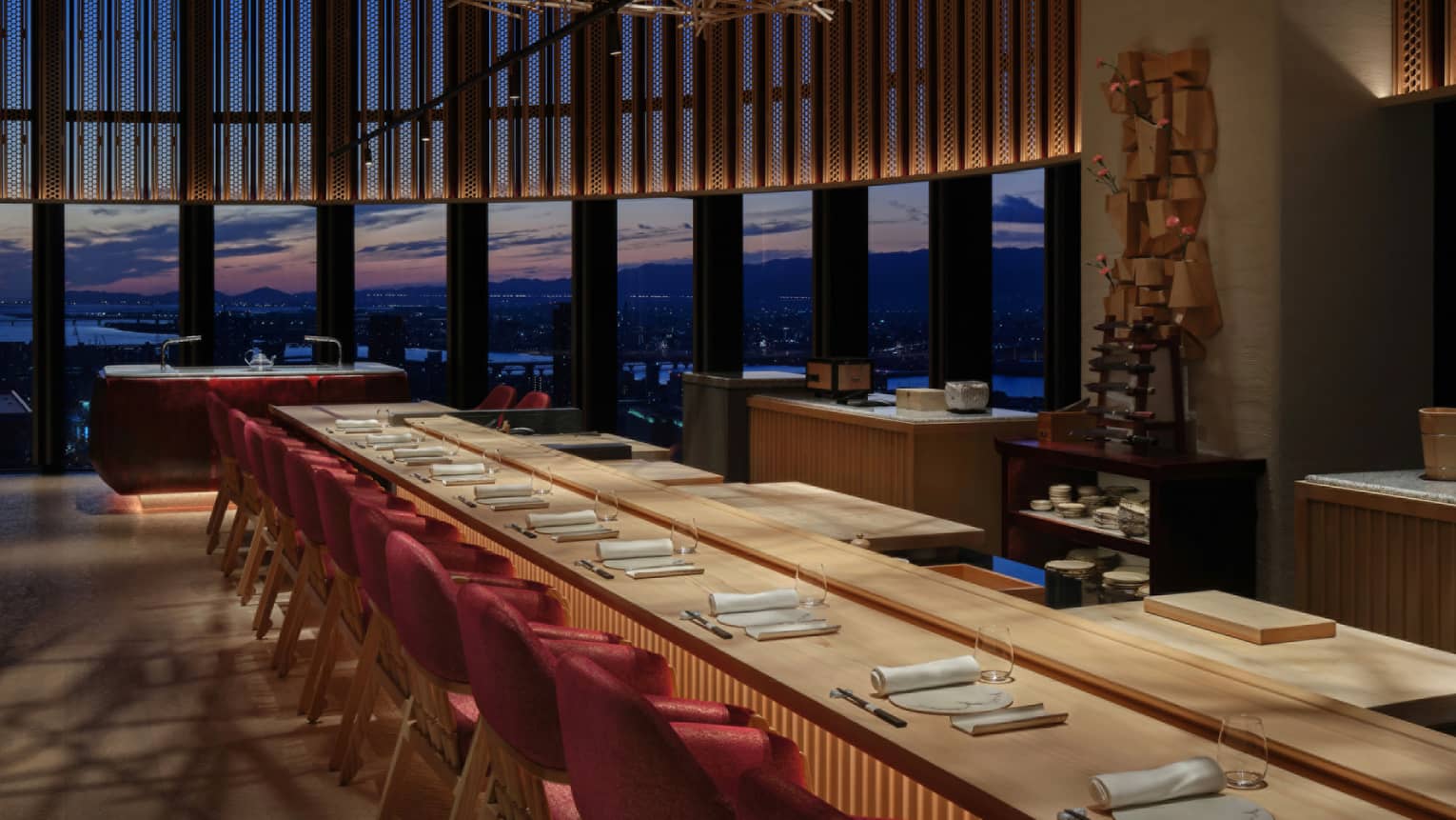 Ten place settings at a long wooden table with warm underlighting, a curved bank of windows offering a dramatic sunset view.
