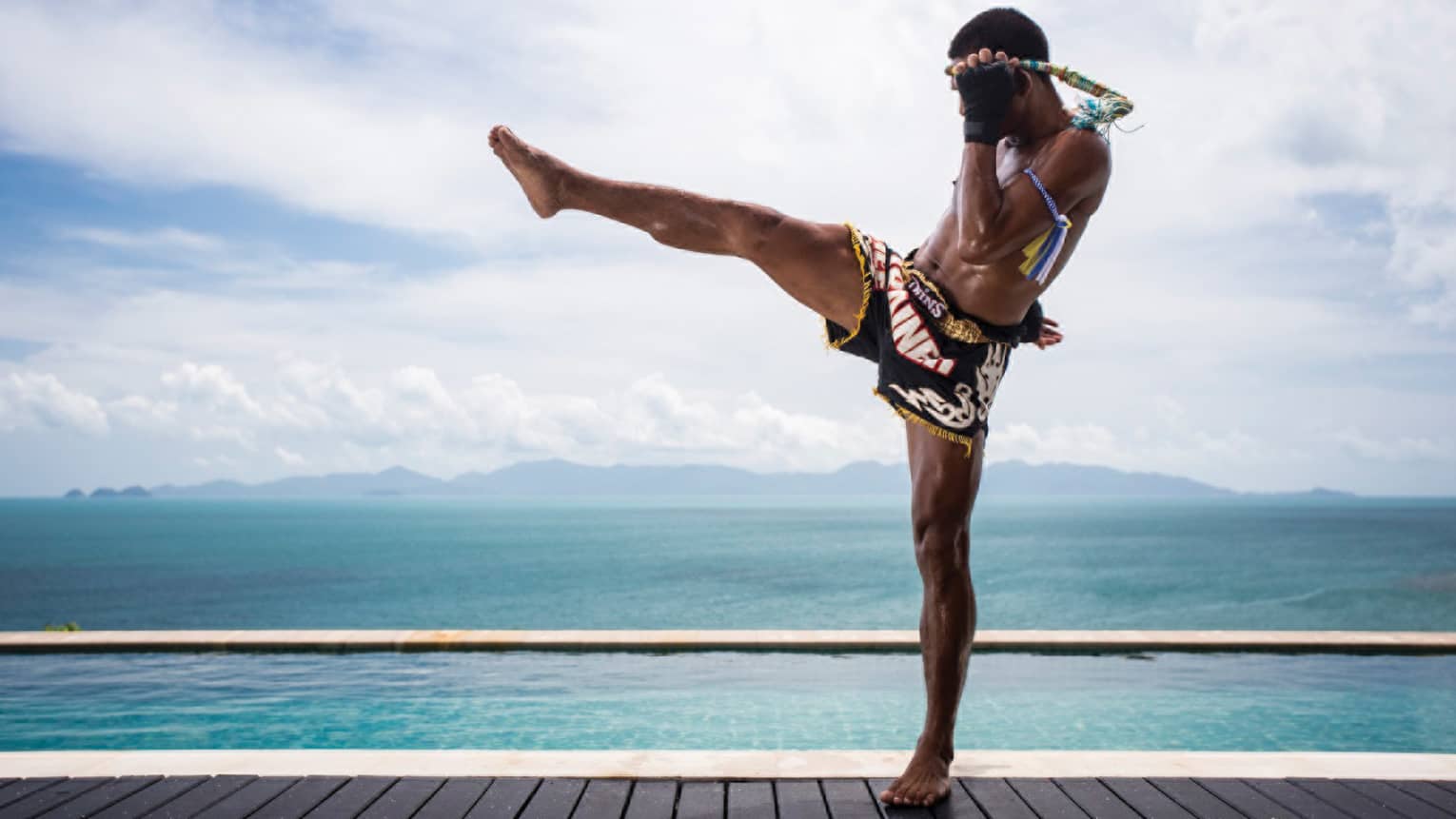 Side view of Muay Thai kick-boxer wearing shorts, doing leg kick on swimming pool deck in front of ocean