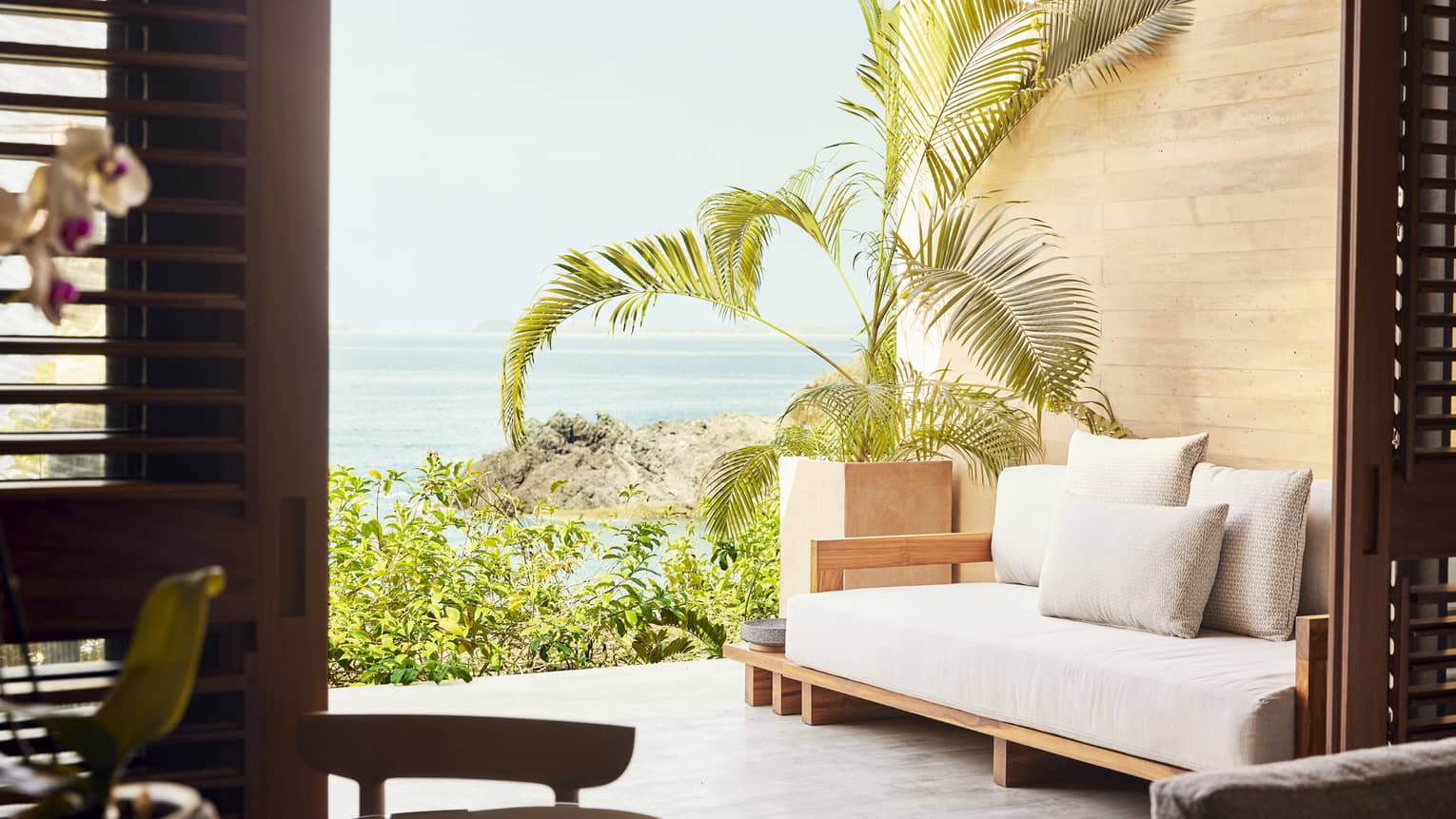 Terrace with a wooden daybed and lush potted plants, offering a view of the ocean and rocky coastline. The space is framed by open wooden shutters.