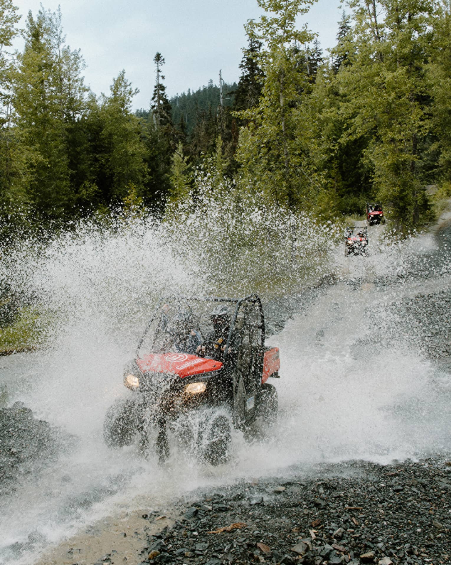 A red-and-black ATV driving through water, creating a thick veil of water spray against a background of pine forest.