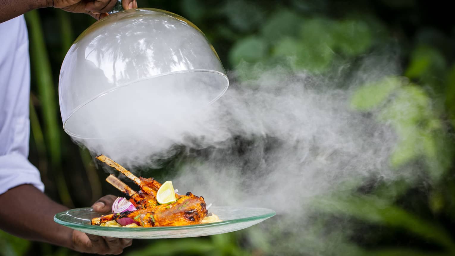 A Four Seasons dining staff taking the lid off of a dinner tray – steam is pouring out over a cooked meat dish on skewers