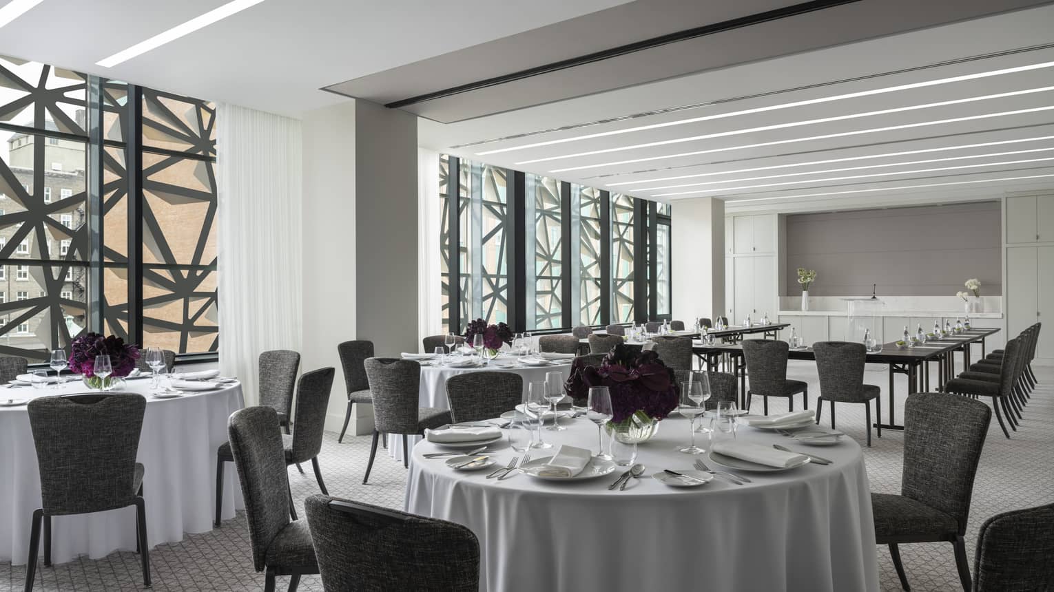 Salon with white round tables and flowers, grey chairs, U shaped table in background, ceiling-to-wall windows