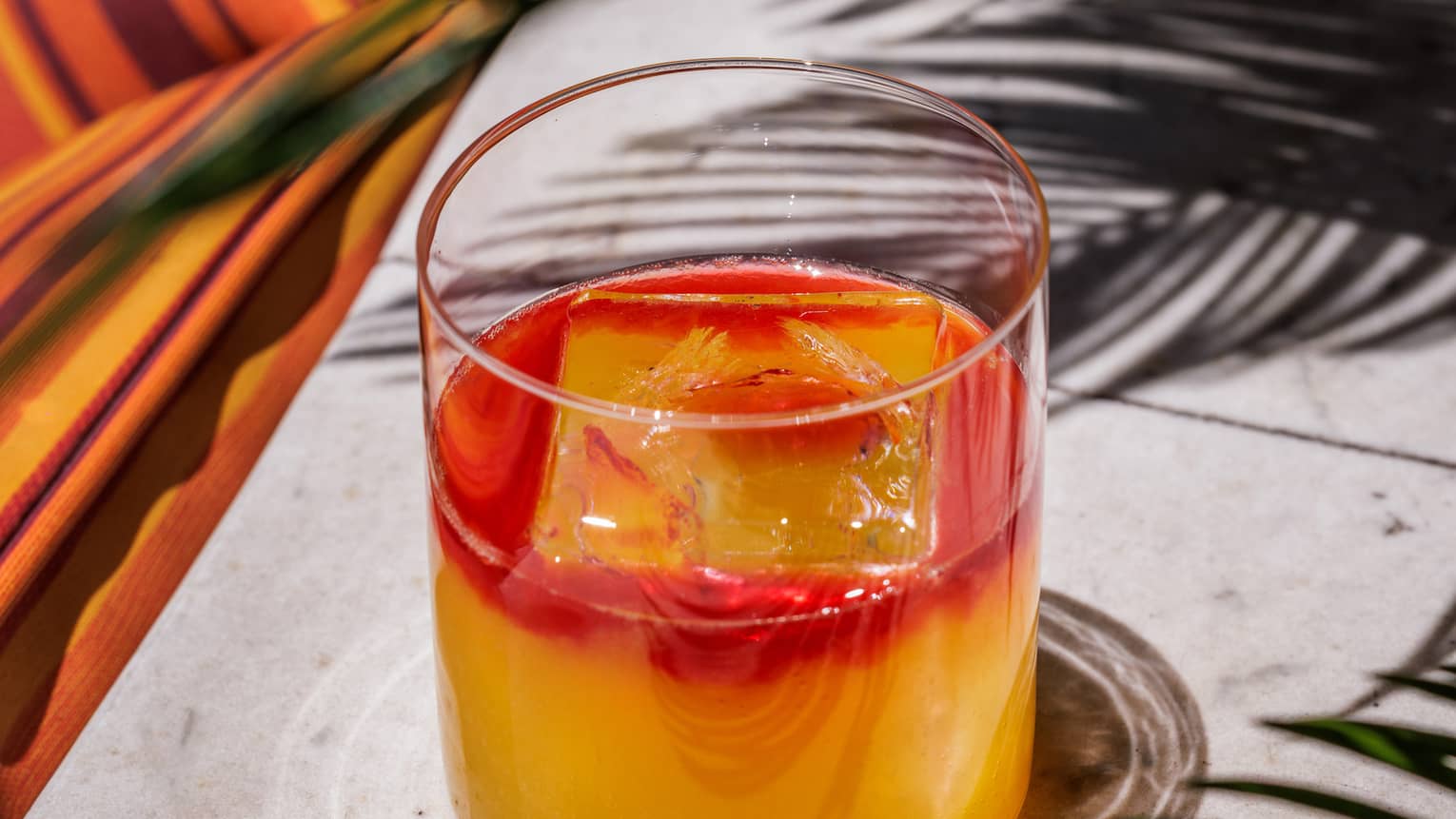 Peach-and-red-coloured mocktail in a clear rocks glass set on a stone table next to palm fronds