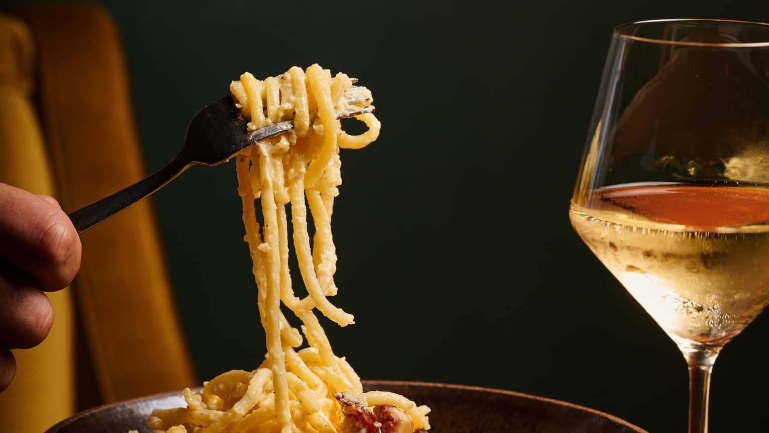Close-up of a fork lifting spaghetti in a bowl of rich, creamy carbonara sauce with a glass of white wine to the right.