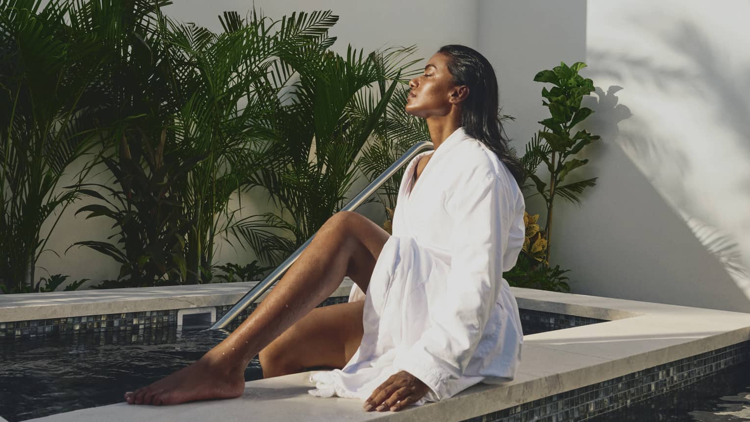 A guest in a linen bathrobe soaks in the sunlight while dipping their feet in a tiled pool surrounded by walls and plants.