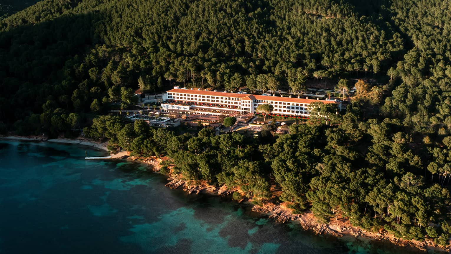 An aerial view of a wooded, mountainous coastline with a resort building among the trees, blue water in the foreground, and mountain views and blue sky in the background
