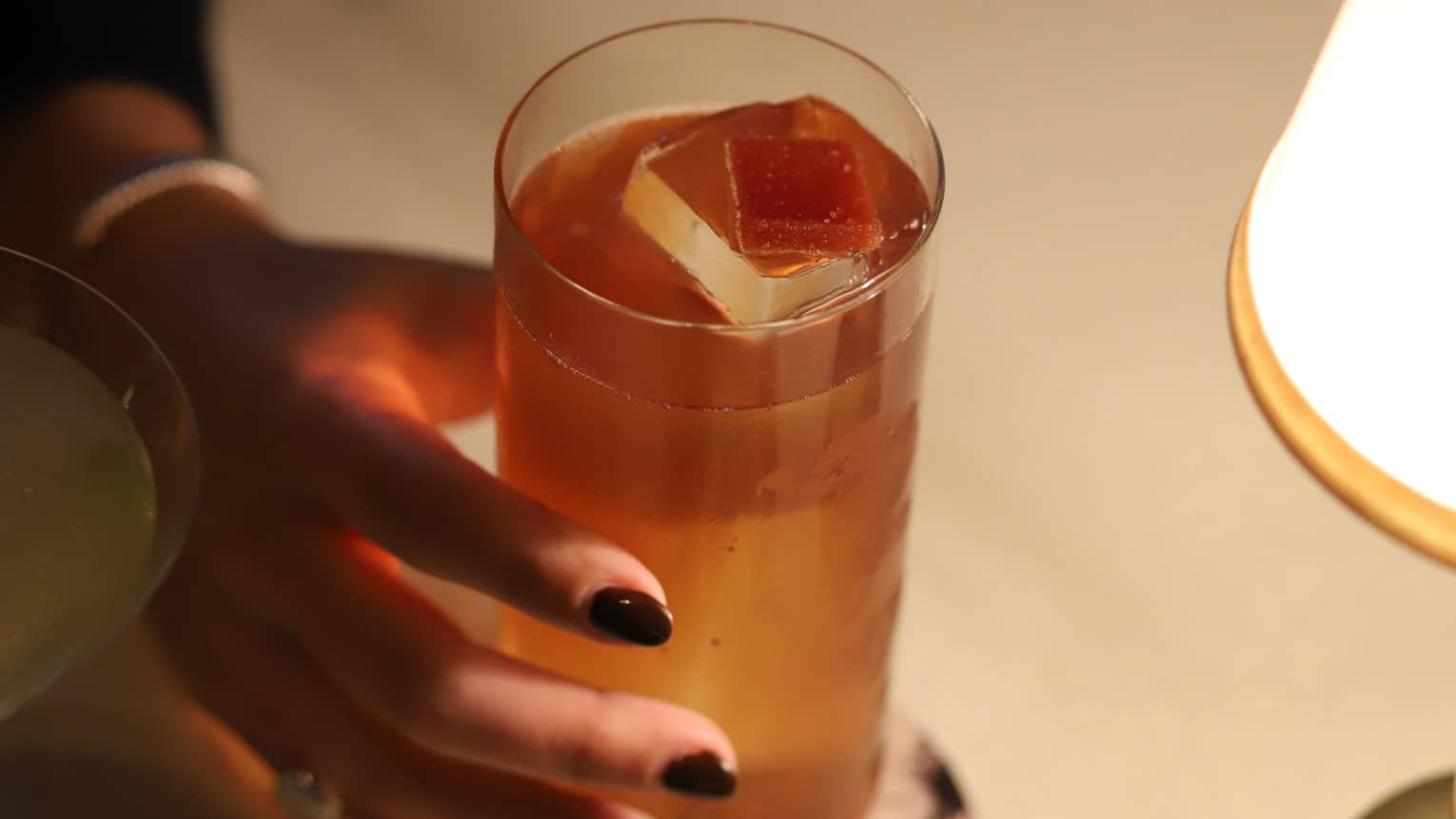 A person holding a glass with a large ice cube in it that is placed on a coaster