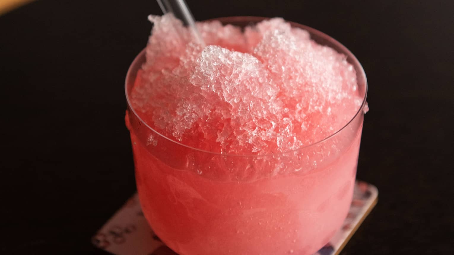 Bright-pink rhubarb ice with clear glass straw in old-fashioned glass atop coaster
