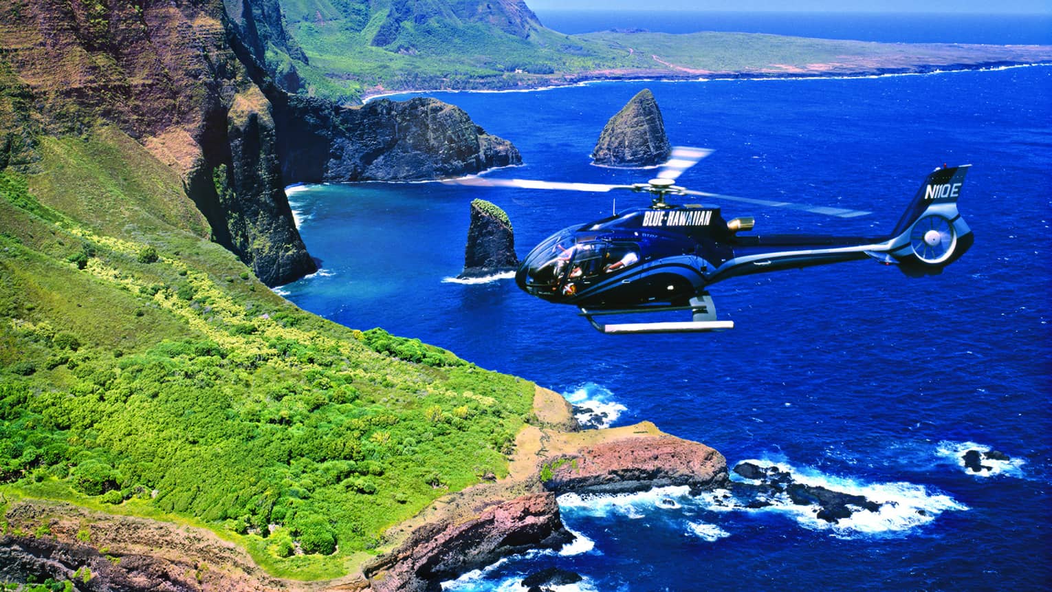 Helicopter flies over ocean and sweeping green cliffs 