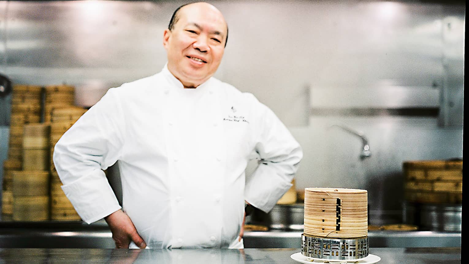 Chef Chan Yan Tak stands smiling in a professional kitchen with his hands on his hips, wearing a white uniform. Bamboo steamers are on the counter. 