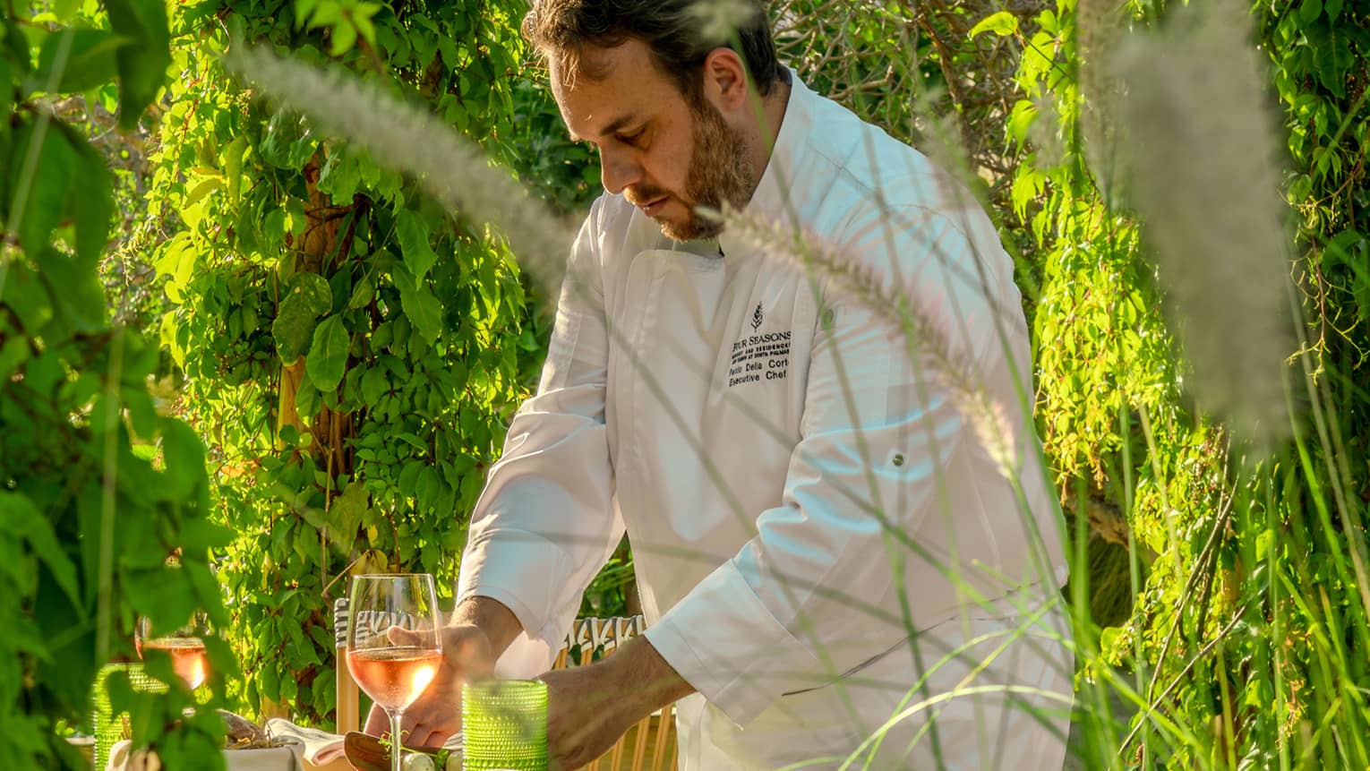 A chef works at a small dining table in a lush, green garden setting.