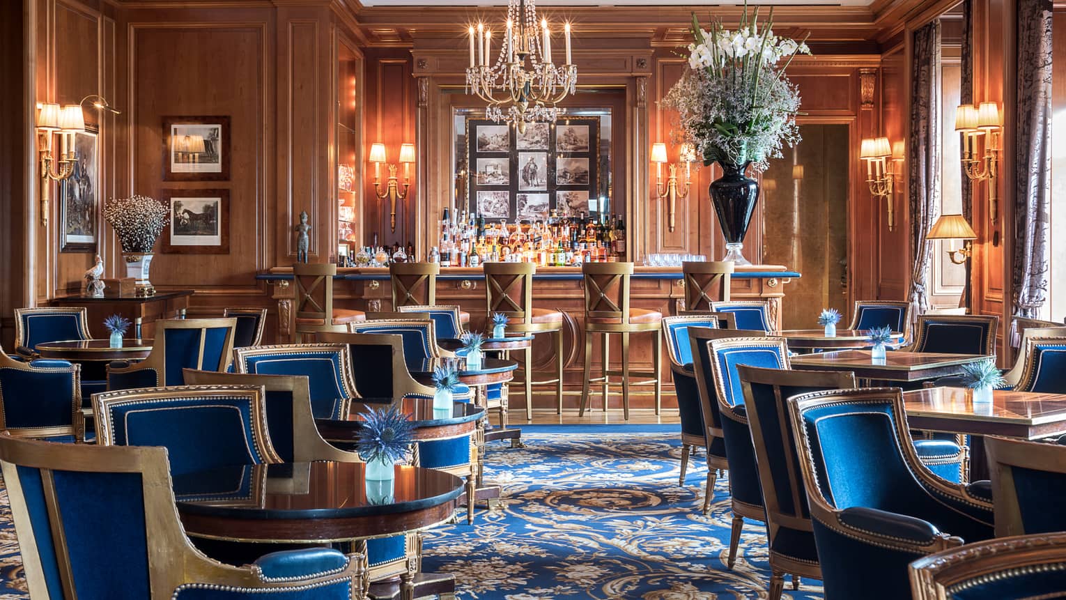 Le Bar des Bergues lounge with elegant blue velvet chairs with wood trims around cocktail tables, crystal chandelier, bar