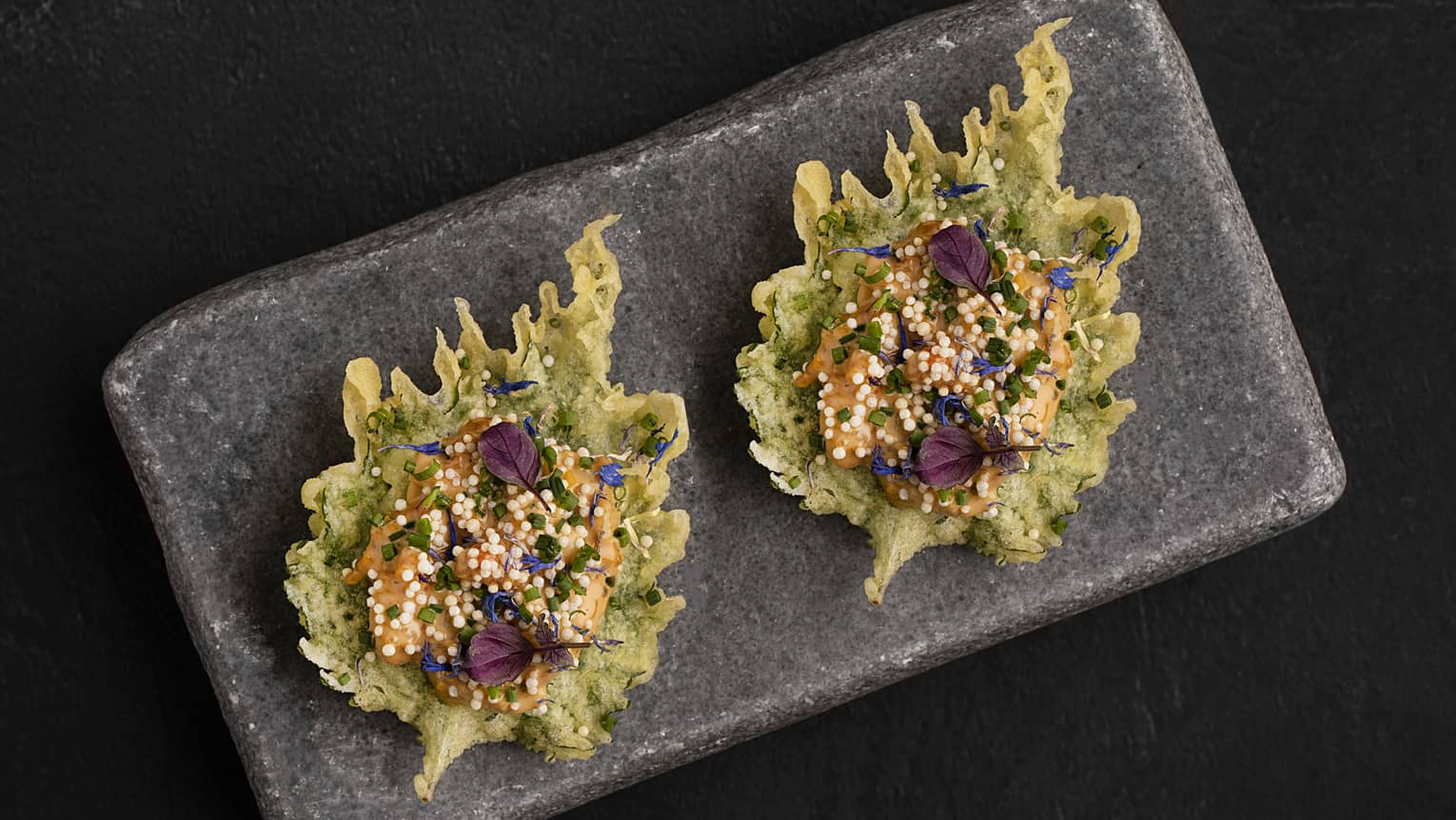 Two green leaves filled with spoonfuls of lobster tartare plates on a rectangular slate-grey dish