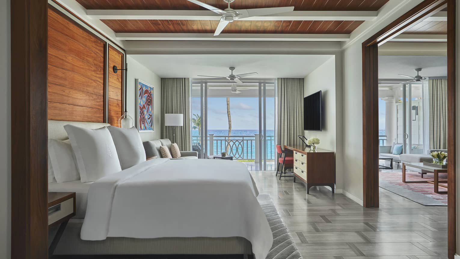 Ocean-view luxury resort bedroom with wooden accents, king bed, seating area, desk, wall-mounted TV and balcony