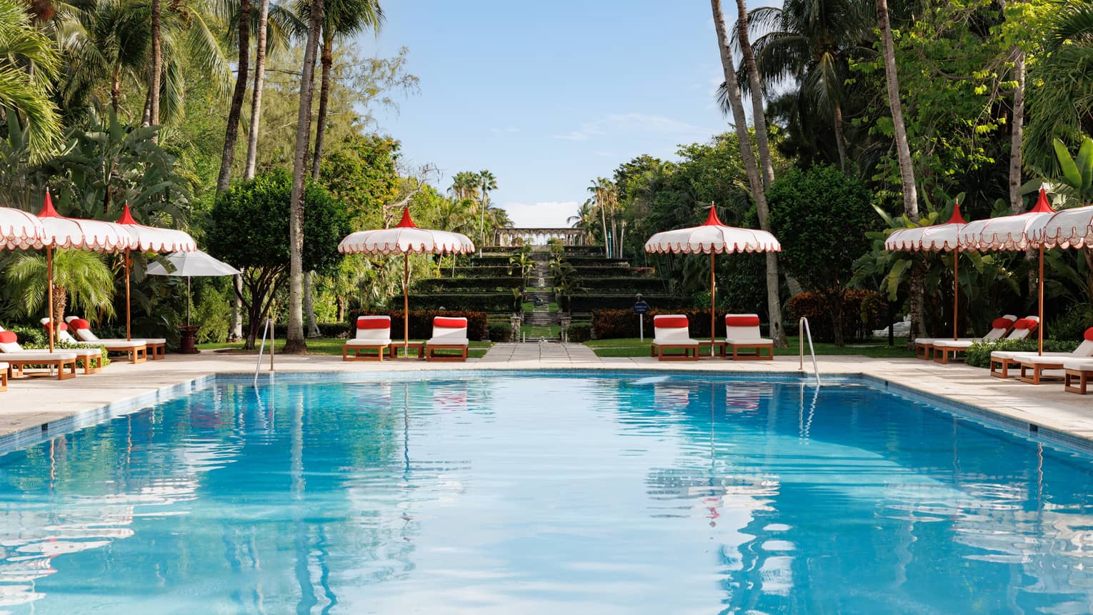 An outdoor pool at a tropical resort, flanked by white and red lounge chairs and umbrellas