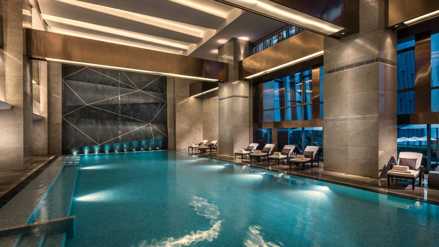 Lounge chairs line indoor swimming pool under marble pillars, ceiling lights