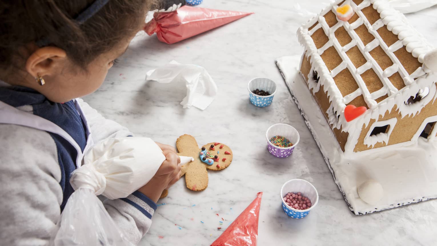 ,Young girl decorates gingerbread man with sprinkles in front of gingerbread house