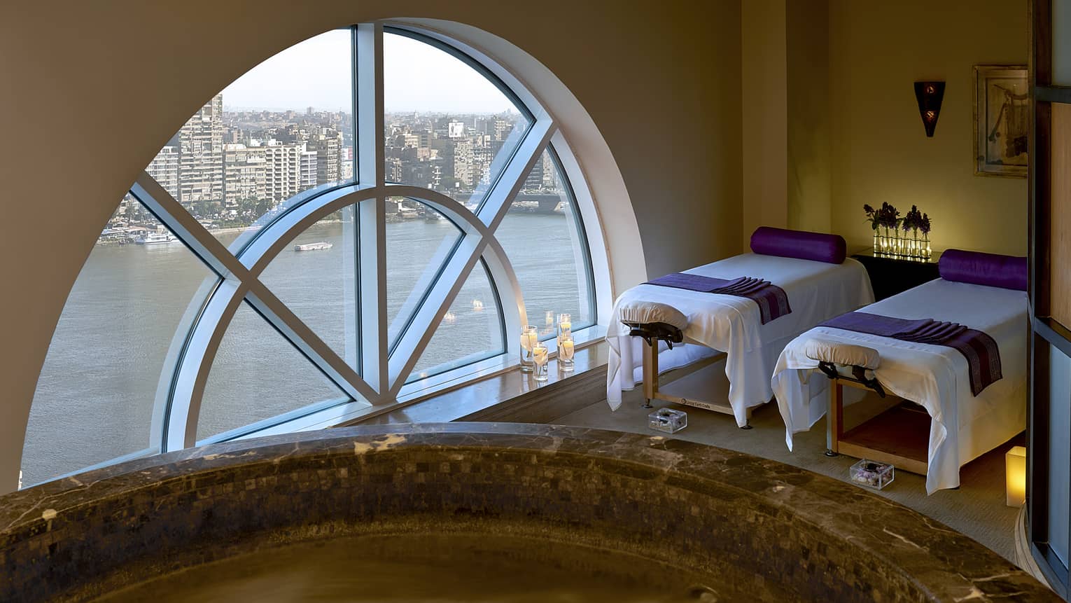 Two massage tables side-by-side in couples spa suite by lare crescent-shaped window overlooking Nile