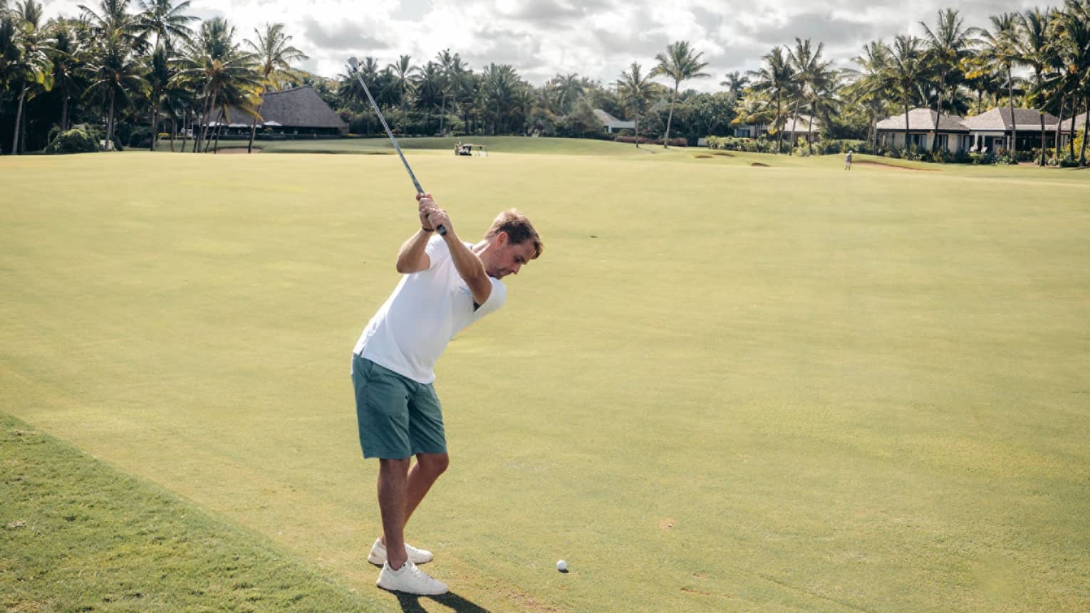 Man in white polo and green shorts swinging a golf club on green with palm trees and cloud-dotted sky in backdrop