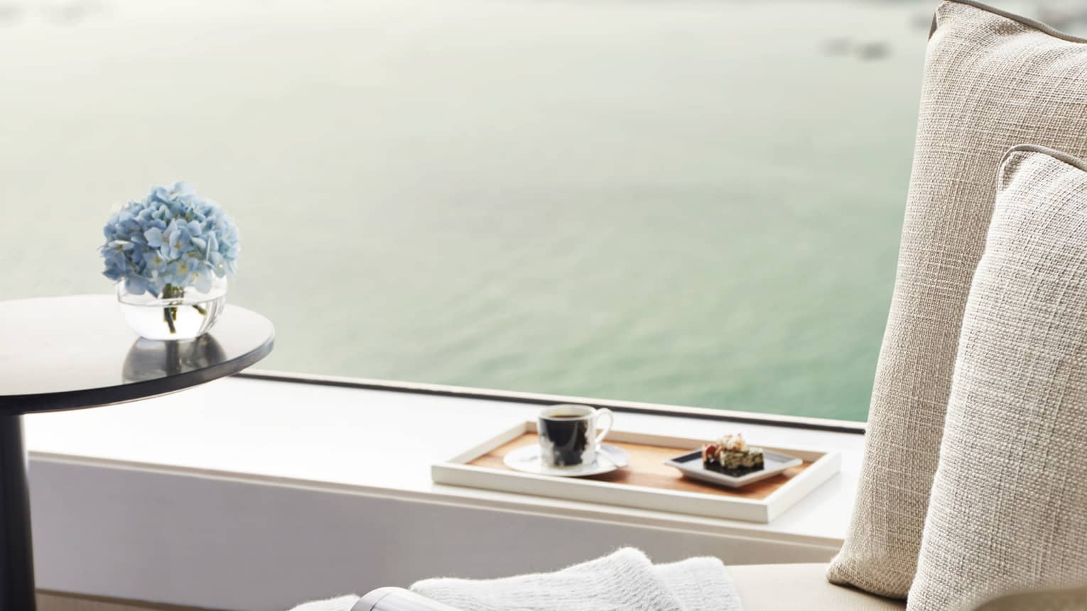 Close-up of cozy seating by window with a cup of coffee, pastries on a tray, a magazine, and blue flowers, overlooking water.