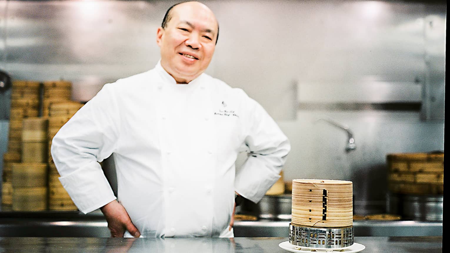 Chef Chan Yan Tak stands smiling in a professional kitchen with his hands on his hips, wearing a white uniform. Bamboo steamers are on the counter. 