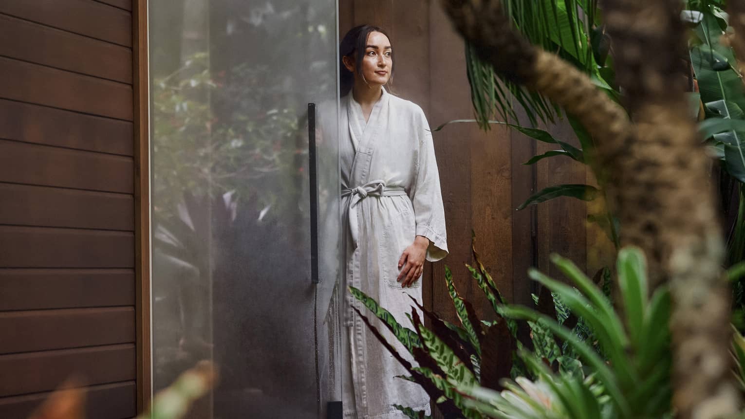 A woman dressed in a bathrobe stands by a glass door at an outdoor spa area surrounded by lush greenery