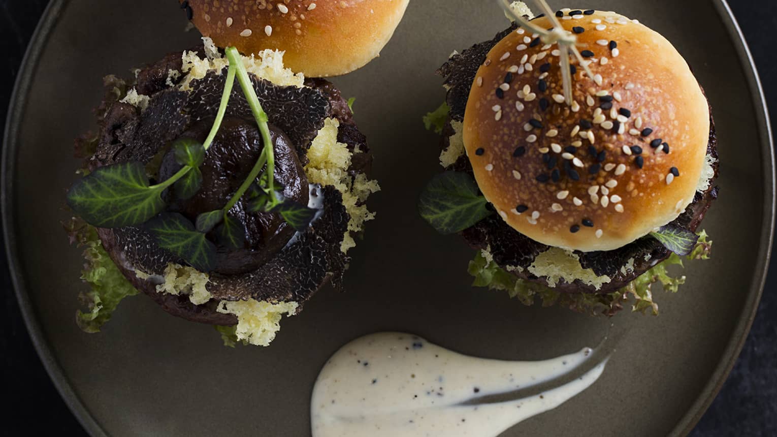 Two Wagyu beef sliders garnished with sesame seeds on a gray ceramic plate with a dollop of a white creamy dressing on the side