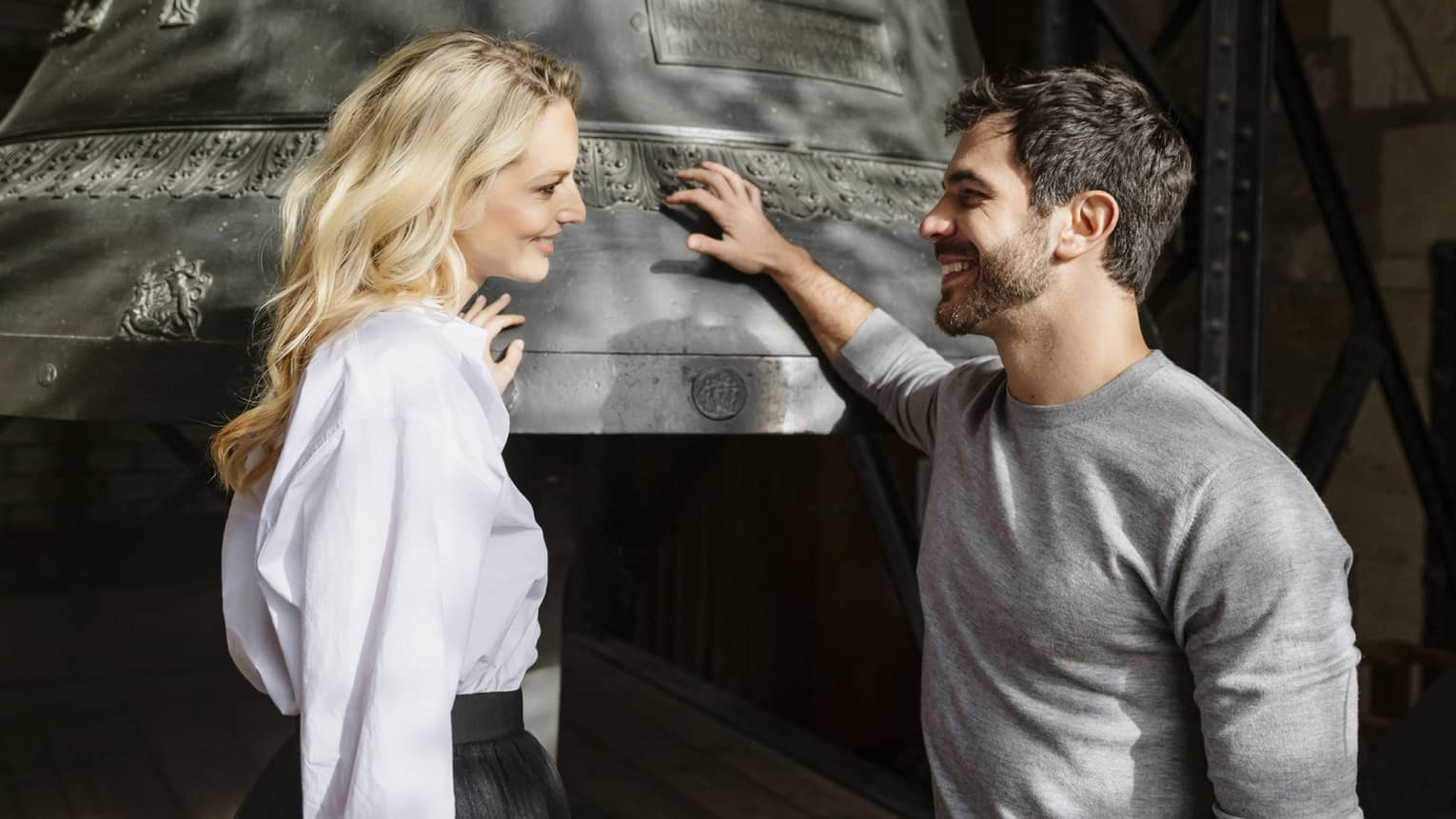 Two people stand next to a large metal bell in the bell tower of a cathedral.