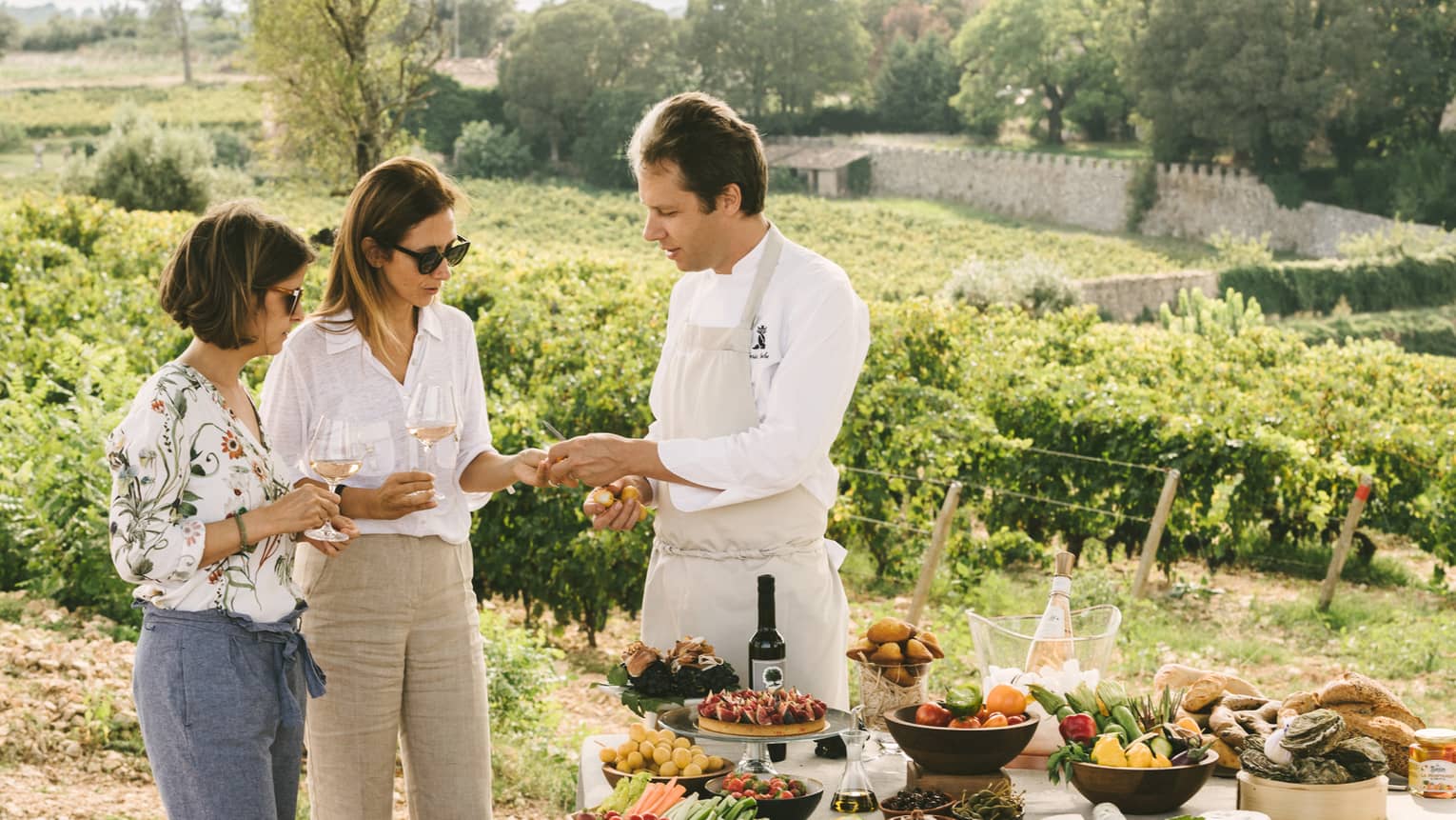 Two women hold wine glasses, stand by chef at outdoor buffet table in garden
