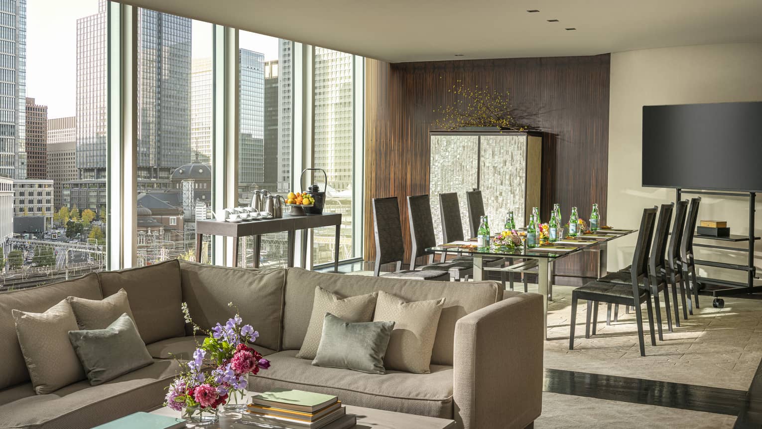 Living area with a large window, city skyline view, beige sectional sofa, table with refreshments, dining area with black chairs and home theatre