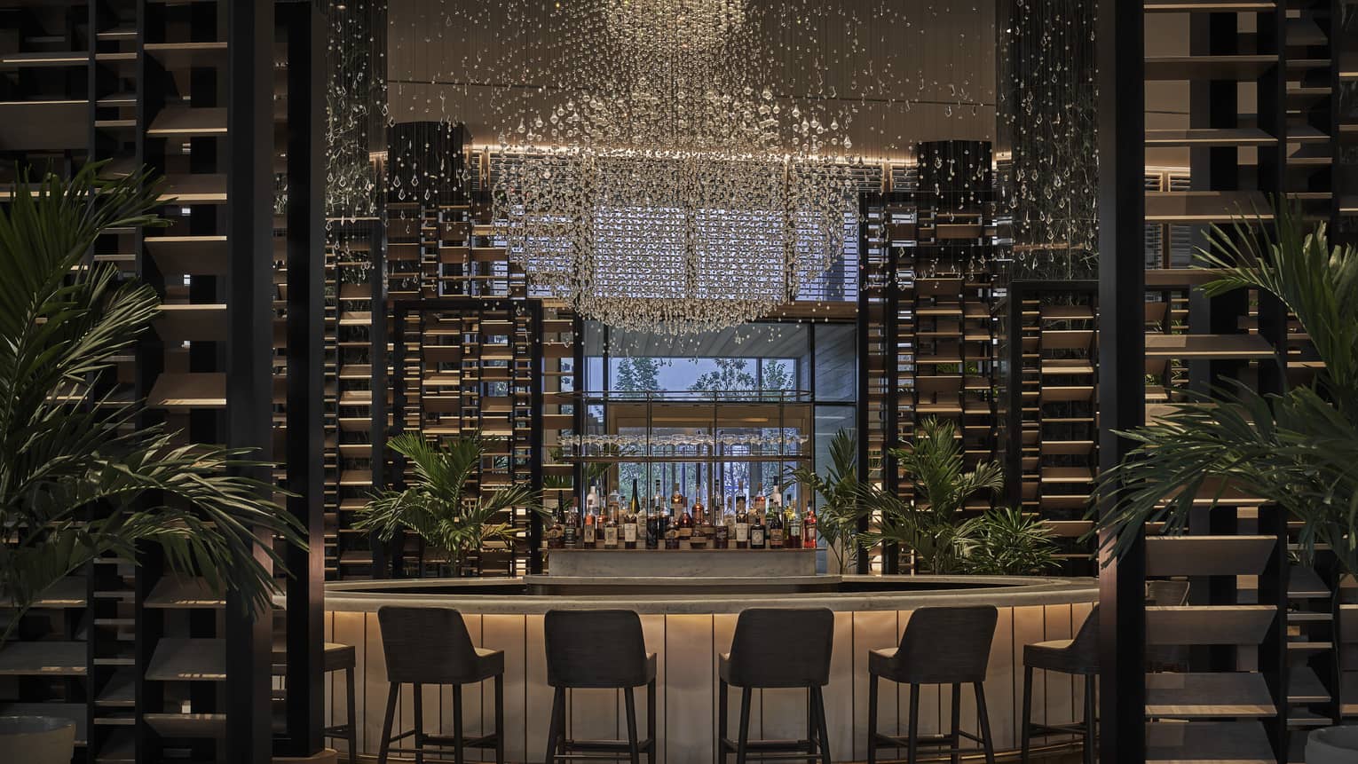 Bar at Chandelier Bar with six stools flanked by ergonomic pillars and palm trees, liquor bottle display in the centre, elaborate chandelier hanging from the ceiling