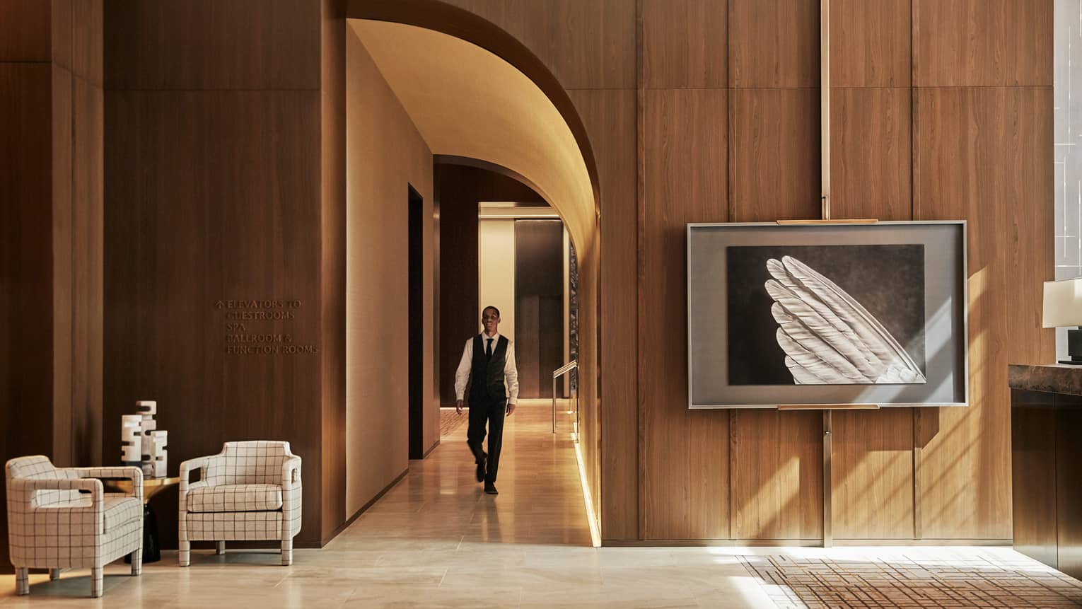 Member of concierge walks down hallway toward hotel lobby, sunlight streaming in from window off camera