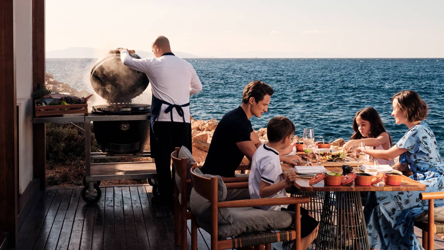 A group of people at an outdoor dining table near the ocean, with the ocean and sky in the background.
