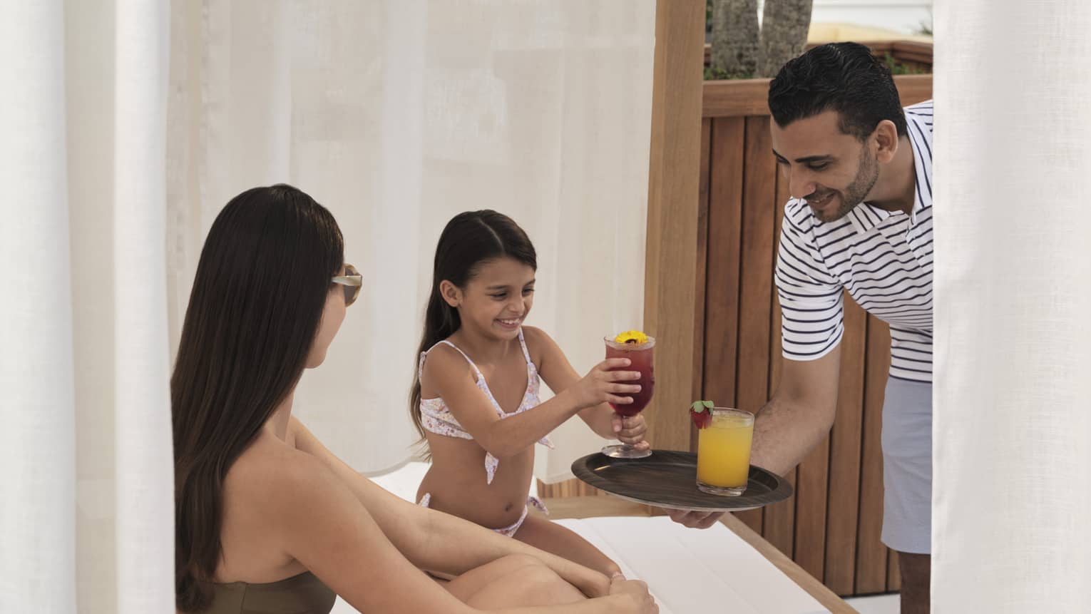 Two guests wearing swimsuits and sitting in a cabana while a person holds a plate with two drinks.