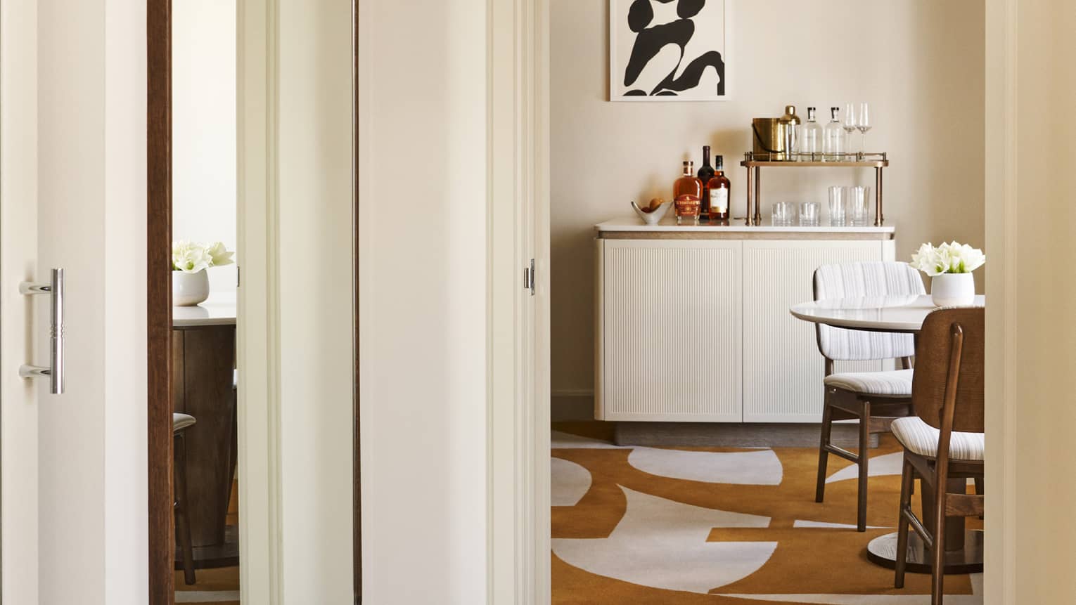 Hotel suite hallway with long mirror on the wall, amber and white abstract carpeting, wet bar and table with two chairs