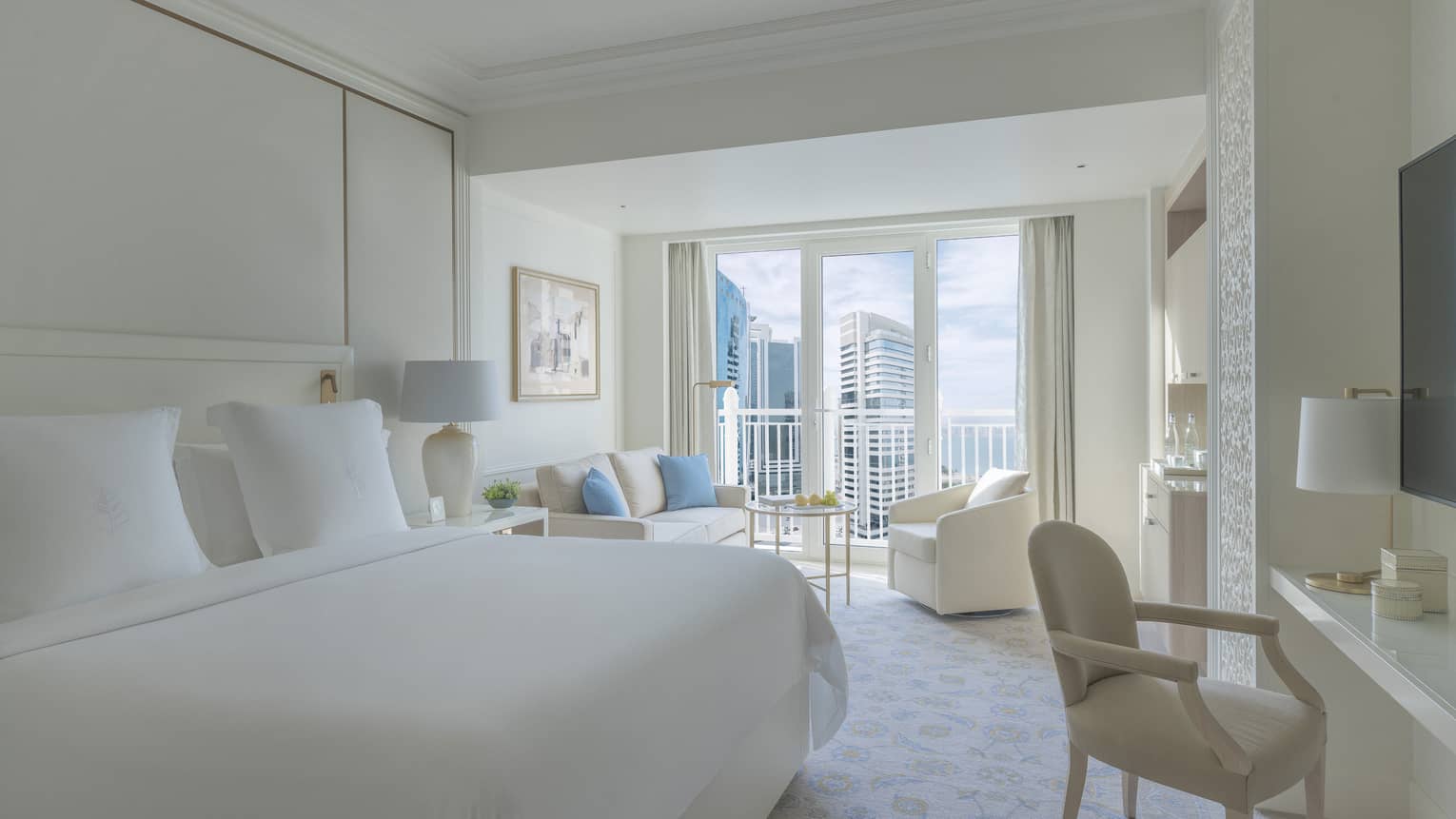 Bedroom with white bed, white walls and a white sofa and arm chair, with balcony offering city view