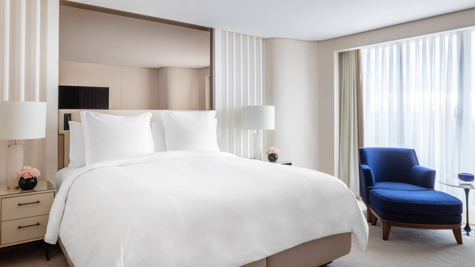 Bedroom with white king bed, royal blue chaise, mirrored headboard, white curtains, nightstands with pink flowers