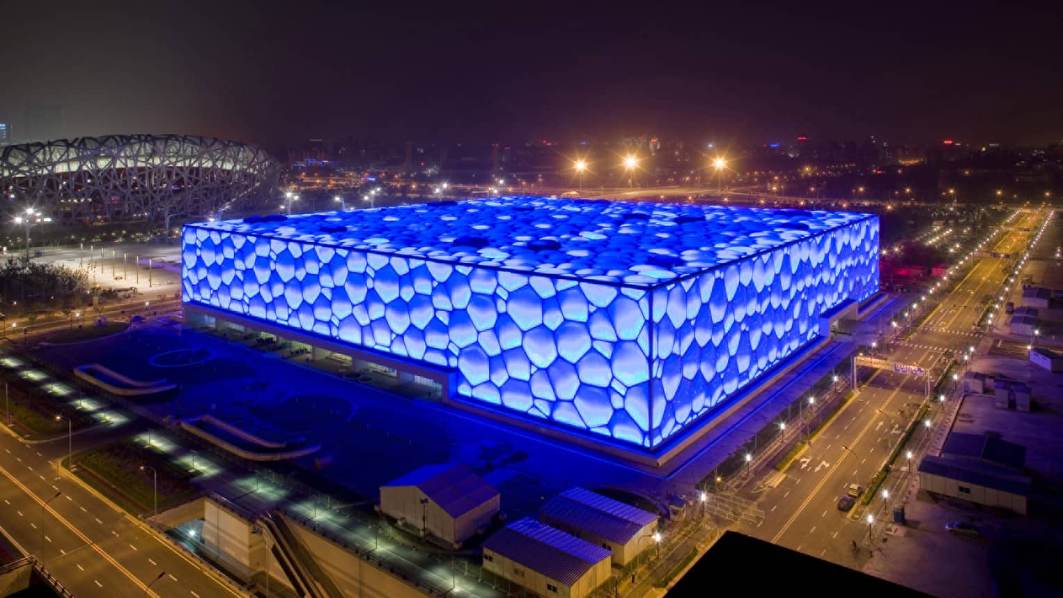 Under a dark night sky, empty city streets border a large cube-shaped building covered in a bright-blue bubble pattern.