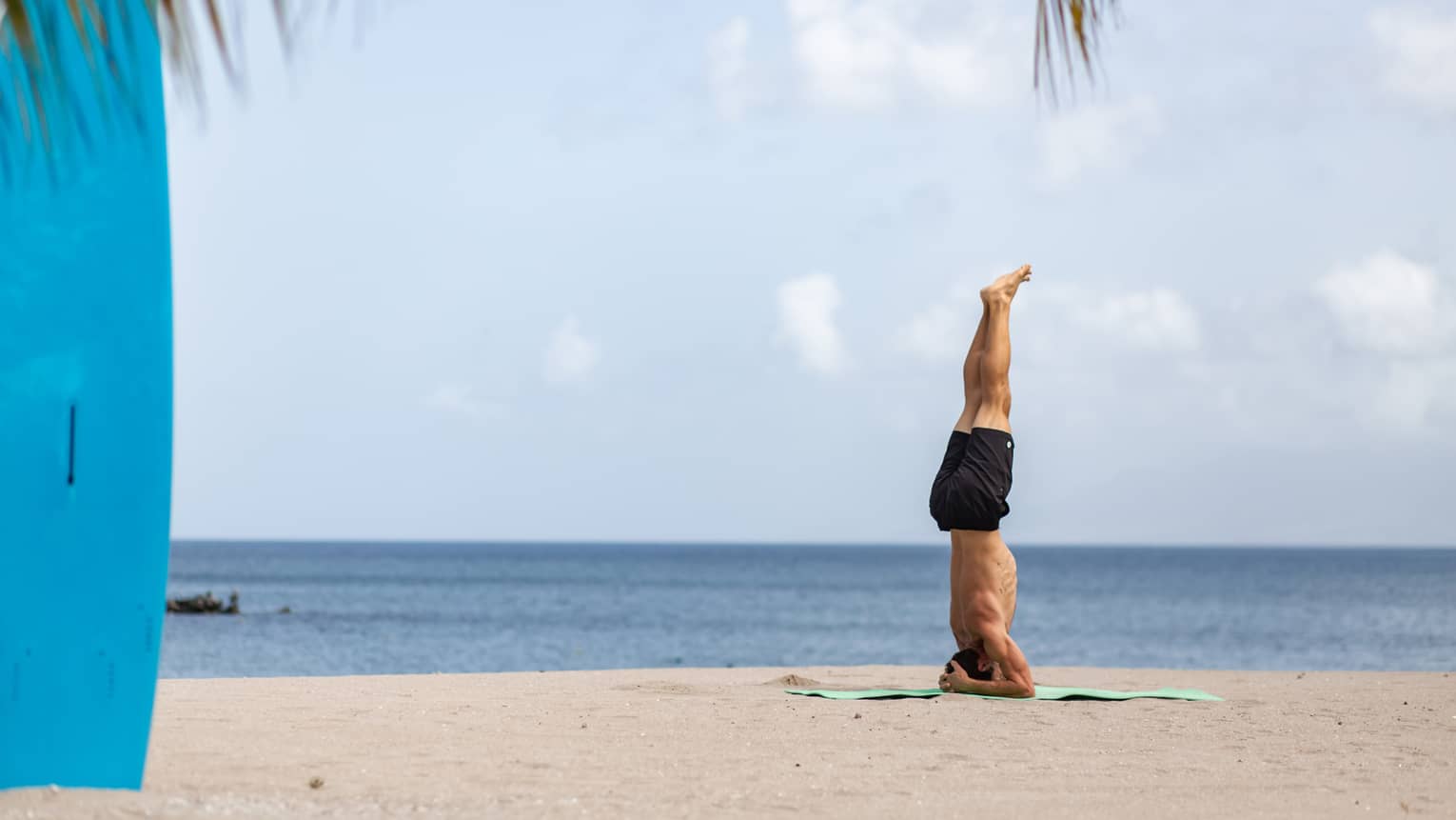 A yogi practices their headstand on the beach under clear skies. Nearby, a bright blue surfboard stands upright in the sand.