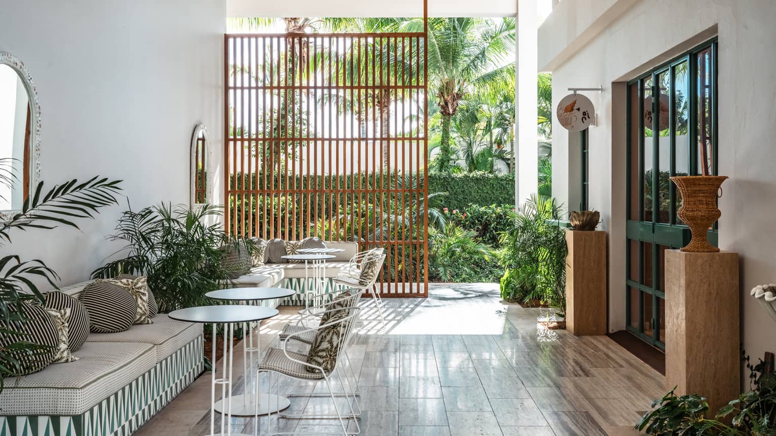 An airy light-filled cafe area with small round tables, bench and chair seating, accents of greenery, and a far wall open to the outdoors