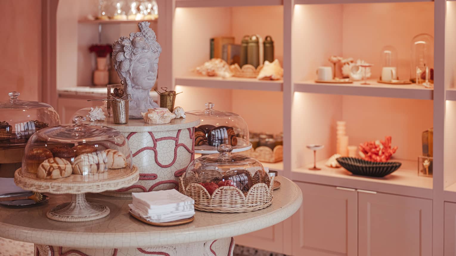 An interior cafe area finished in various shades of pink and white, with display shelves, and a table of food items in decorative platters under glass domes