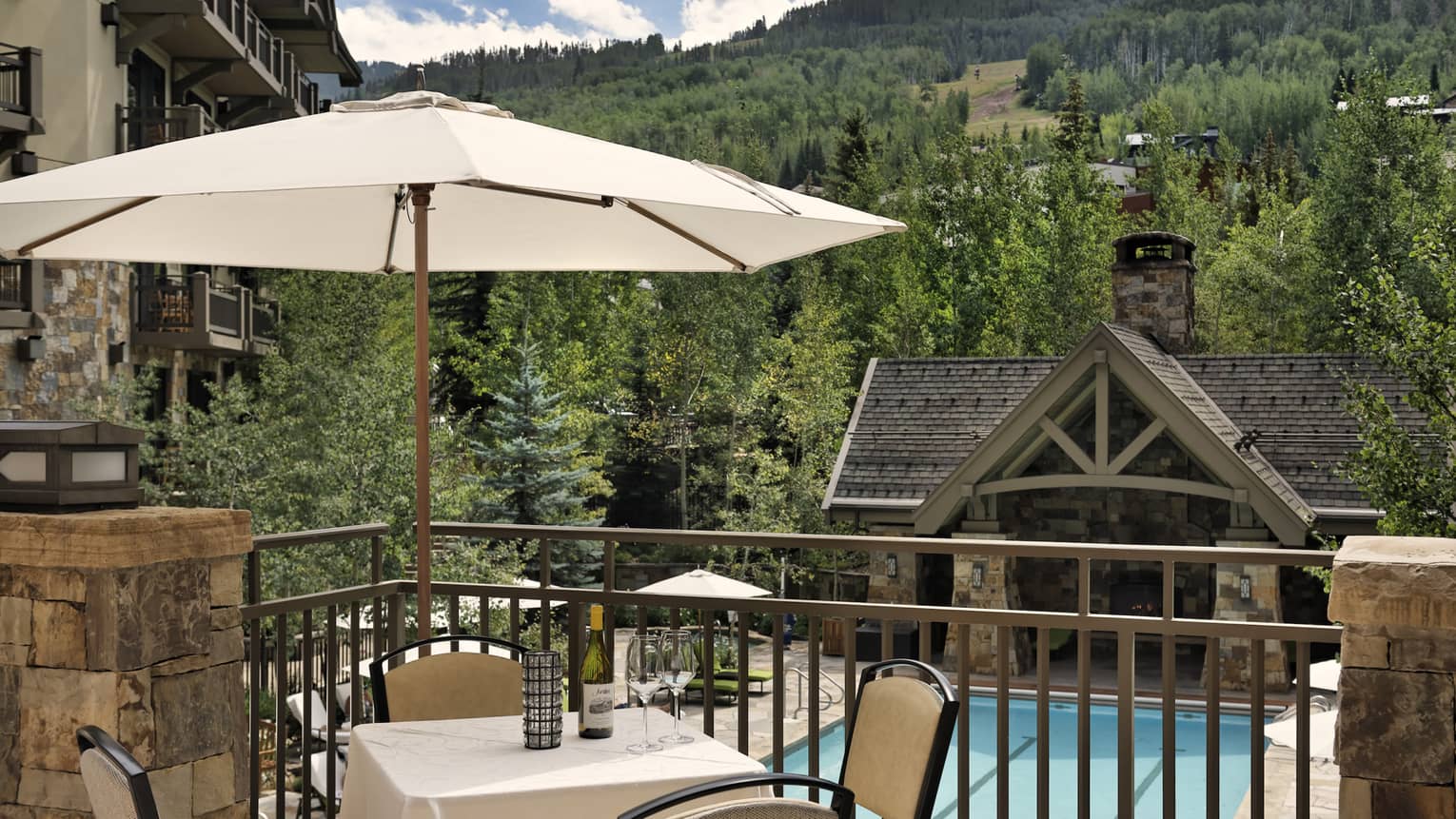 A balcony with a table and umbrella overlooking a pool and a cabin-like building, a forest of trees can be seen in the distance.