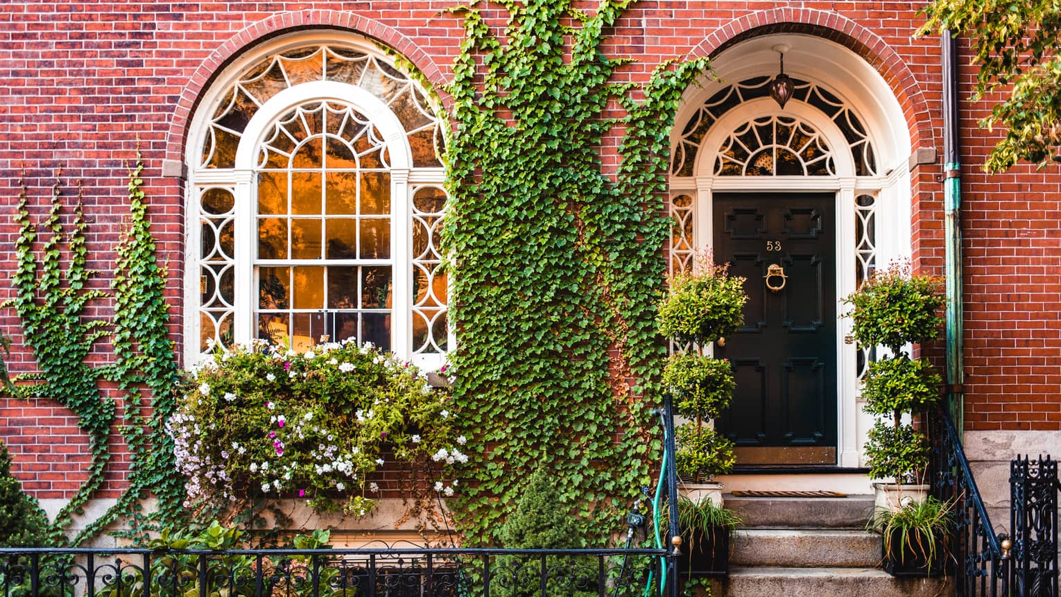 A red brick building with a large arched window, dark door and stairs surrounded by plants.