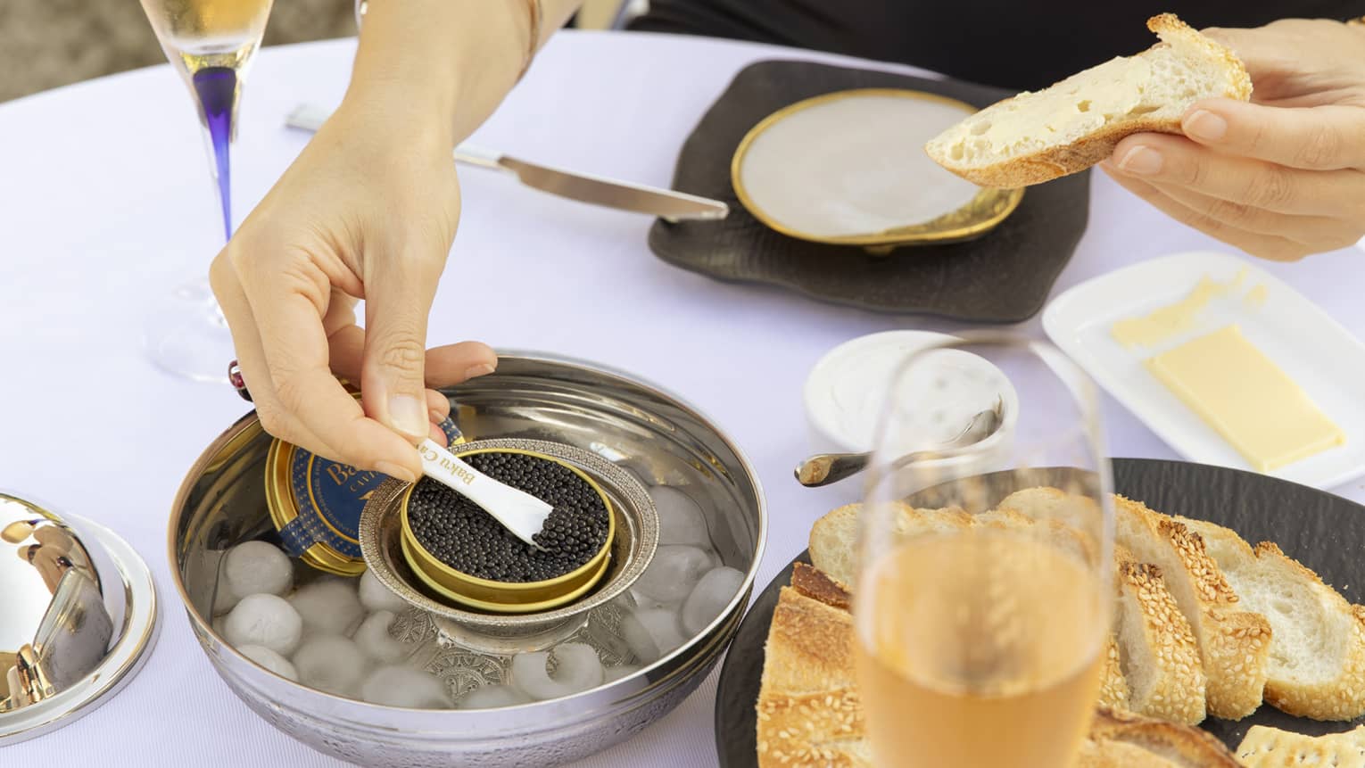 At a table set with champagne flutes, a diner holds buttered bread in one hand and spoons black caviar with the other.