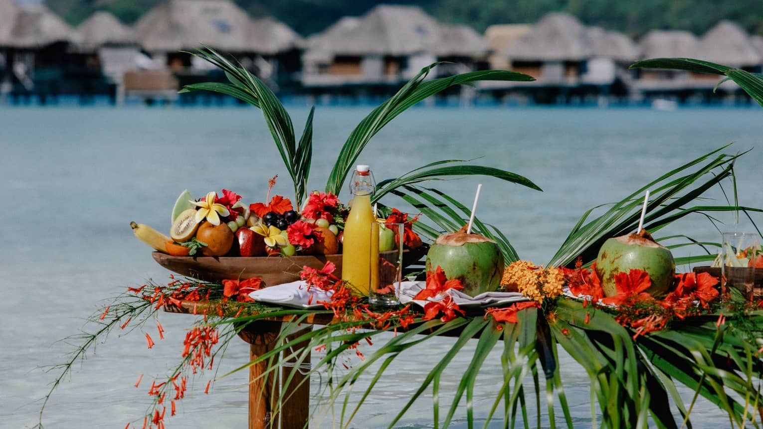 Small wooden table with legs submerged in clear water, topped with assorted fruit and coconuts with straws, mountain and overwater pavilions in background