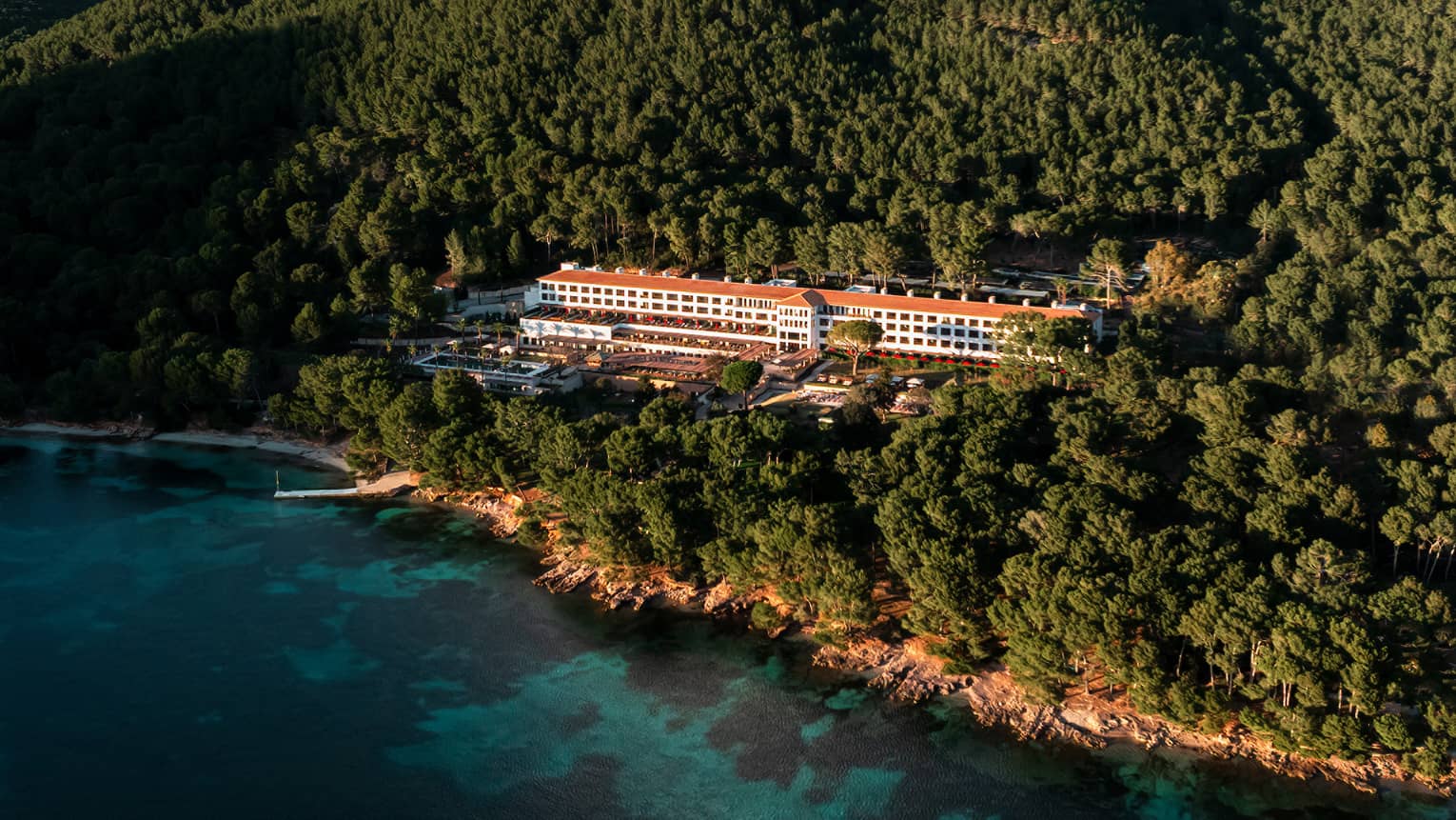An aerial view of a wooded, mountainous coastline with a resort building among the trees, blue water in the foreground, and mountain views and blue sky in the background