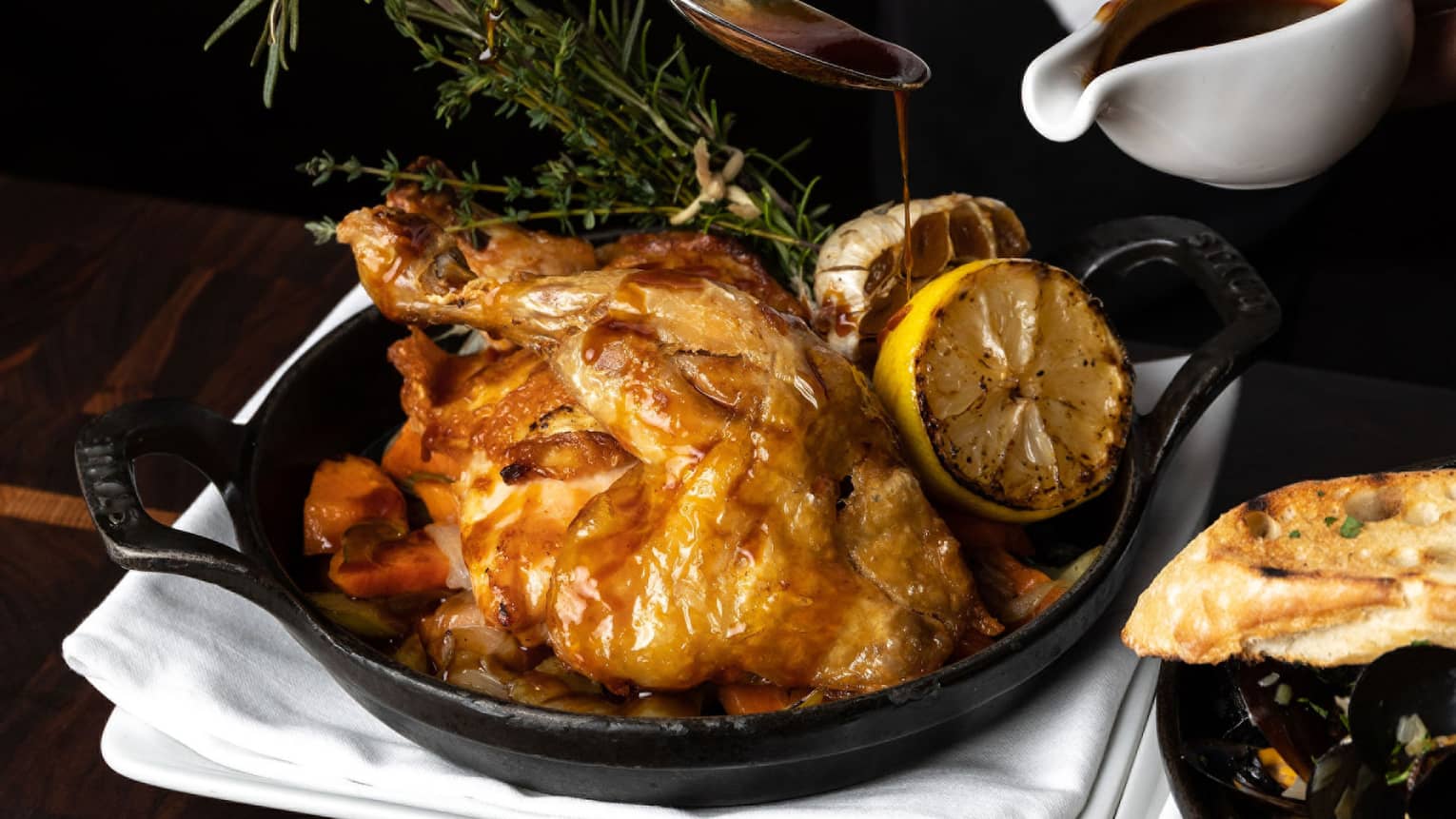 Chef's hands basting a full roasted chicken in an iron skillet, garnished with greenery and roasted lemons